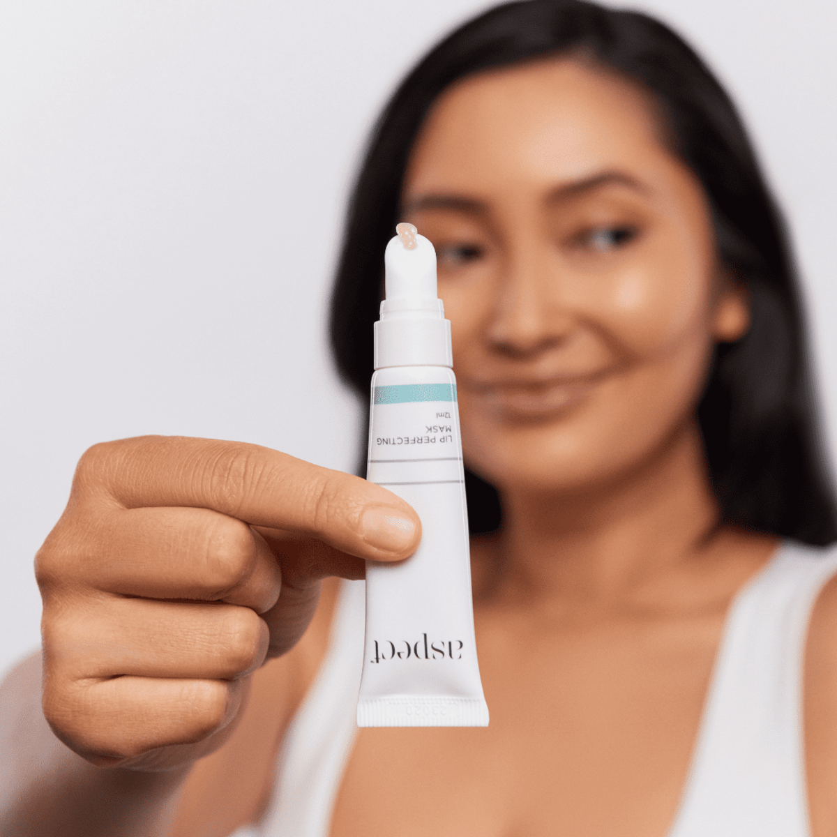 Woman holding Aspect Lip Perfecting Mask with the lid removed and product squeezed out, showcasing the rich, buttery texture that hydrates and plumps lips.